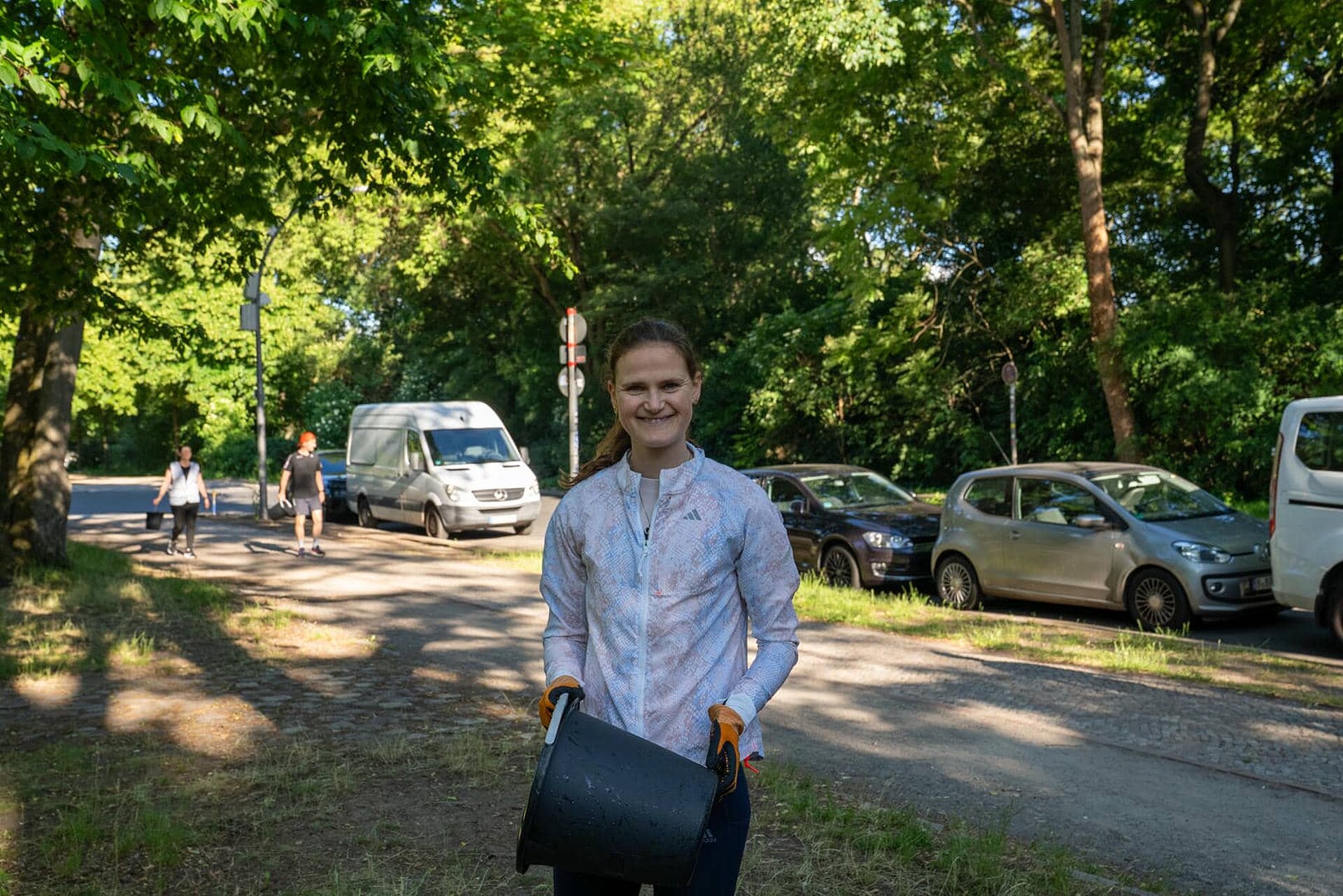Rabea Schöneborn in Arbeitshandschuhen hält einen schwarzen Eimer und lächelt breit. Unscharf im Hintergrund: zwei weitere Teilnehmende mit Eimern.
