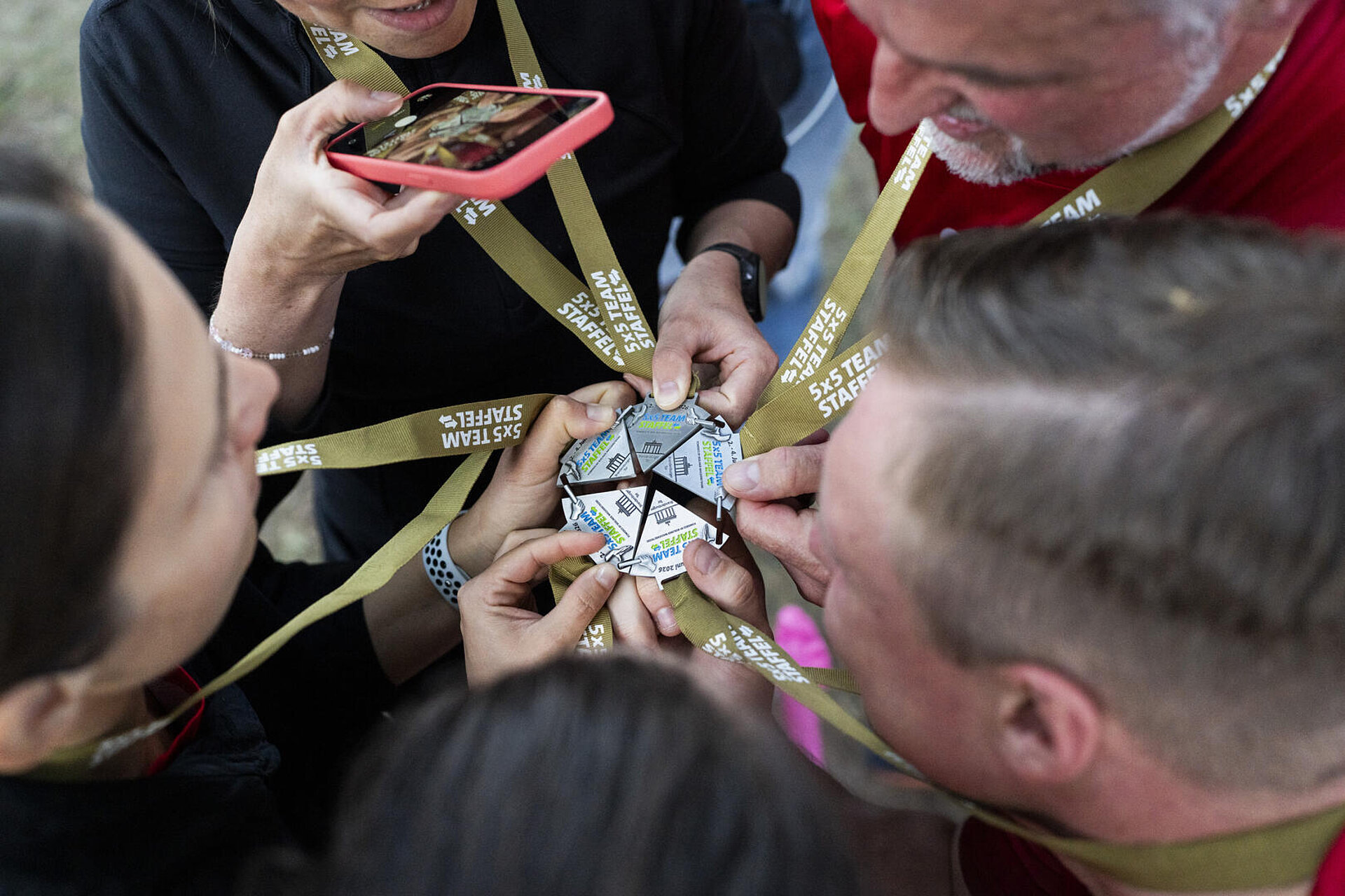5 Teilnehmende setzen ihre Medaillen zur runden Teammedaille zusammen und fotografieren sie. Die Einzelmedaillen sind wie Pizzastücke geformt.