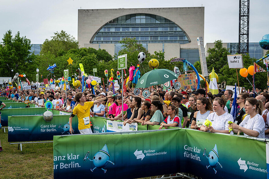 Über der Menschenmenge in der Wechselzone sind zahlreiche Wechselsymbole zu sehen, von Luftballons über Schilder und T-Shirts an Stangen bis hin zu einem Regenschirm.